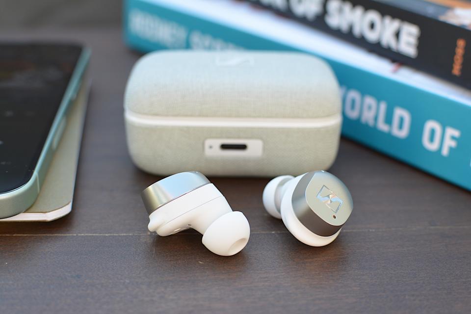<p>White/silver Sennheiser Momentum True Wireless 4 earbuds laying on a wooden table with the case behind them.</p>
