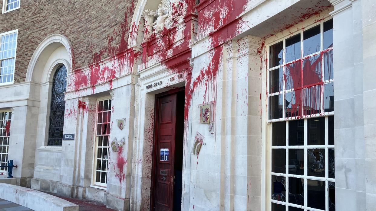 Photo of the front of Somerset Council's building, covered in red paint, with smashed plaques and windows