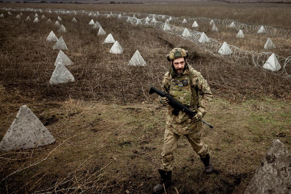 A Ukrainian soldier holds a gun as he walks through a brown field with "Dragon's teeth" concrete triangles and barbed wire behind him