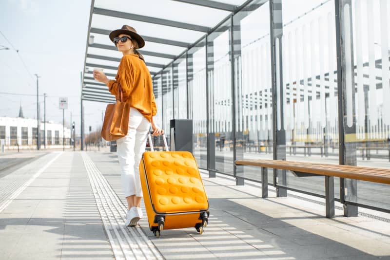 Young female traveler walking with a yellow suitcase at the modern transport stop outdoors, back view. Concept of an urban transportation and travel