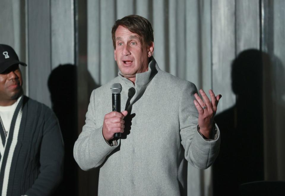 bryan freedman, wearing a gray wool coat, speaks into a microphone on a stage with a gray curtain in the background