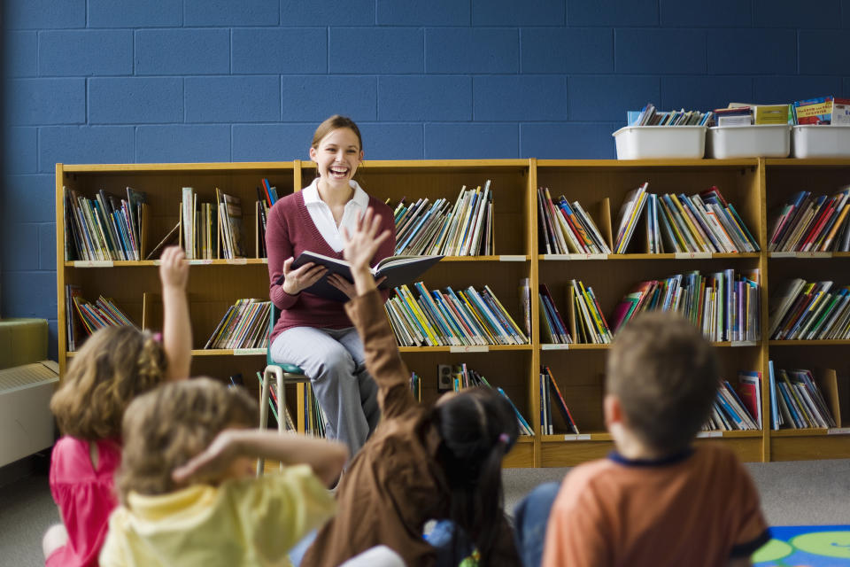 A teacher is sitting on a stool, reading to a group of young children who are sitting on the floor with their hands raised in a classroom library