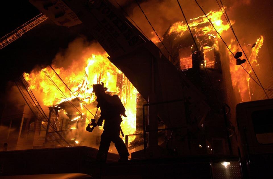 A Yonkers firefighter crosses through the fire scene, with 98 Oak Street fully engulfed in flames behind him on March 14, 2003. The Pre-War wooden buildings with wooden porches and balconies were packed tight together and fire leapfrogged across Nodine Hill in no time.