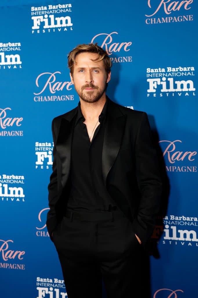 ryan gosling looks at the camera while standing in front of a blue background with written logos on it, he wears all black and a chain necklace