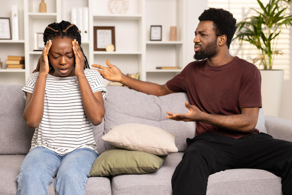 A woman looks distressed while sitting on a couch, holding her head as a man beside her gestures as if trying to explain something