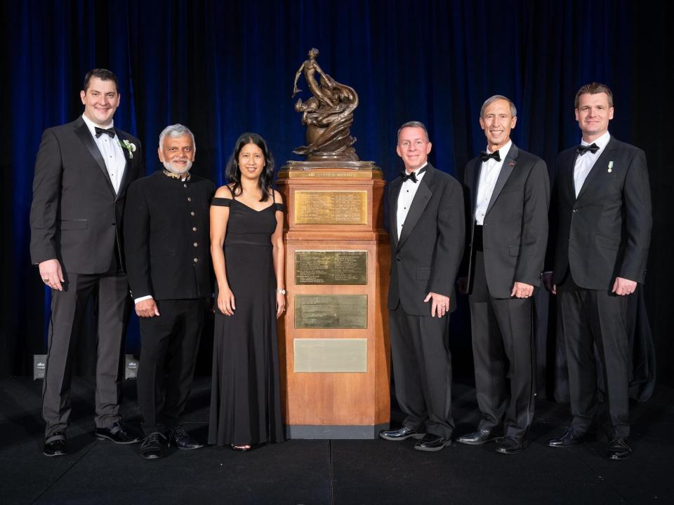 six people in formal dress stand around a human-sized trophy