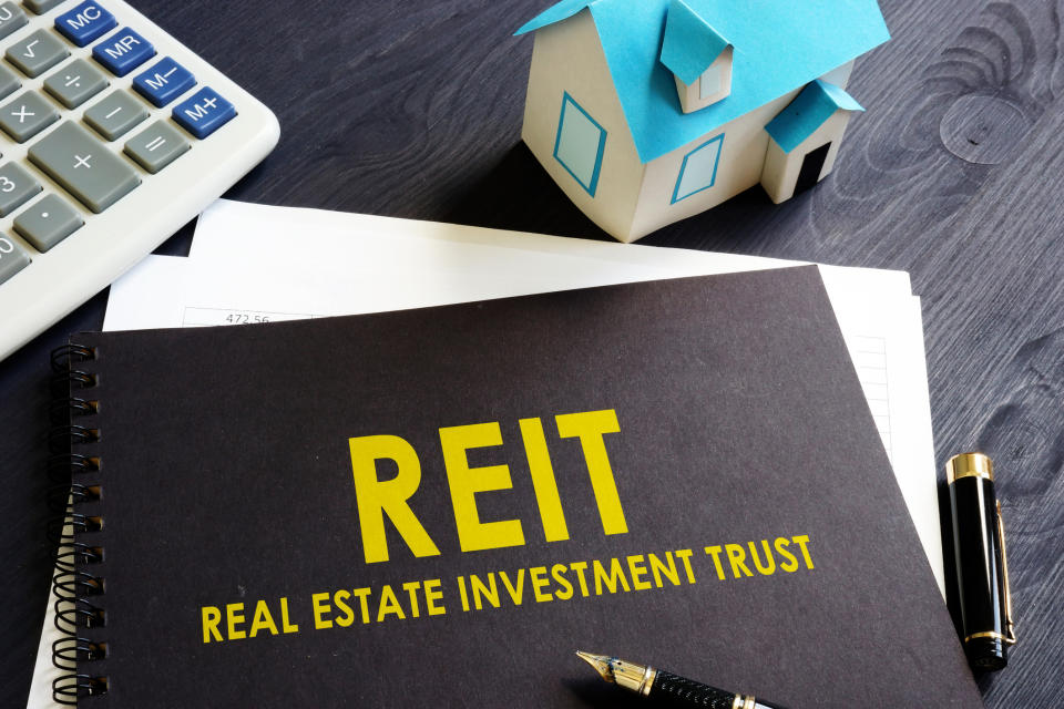 The acronym REIT and the words real estate investment trust on a binder, next to a miniature house, a pen, and a calculator