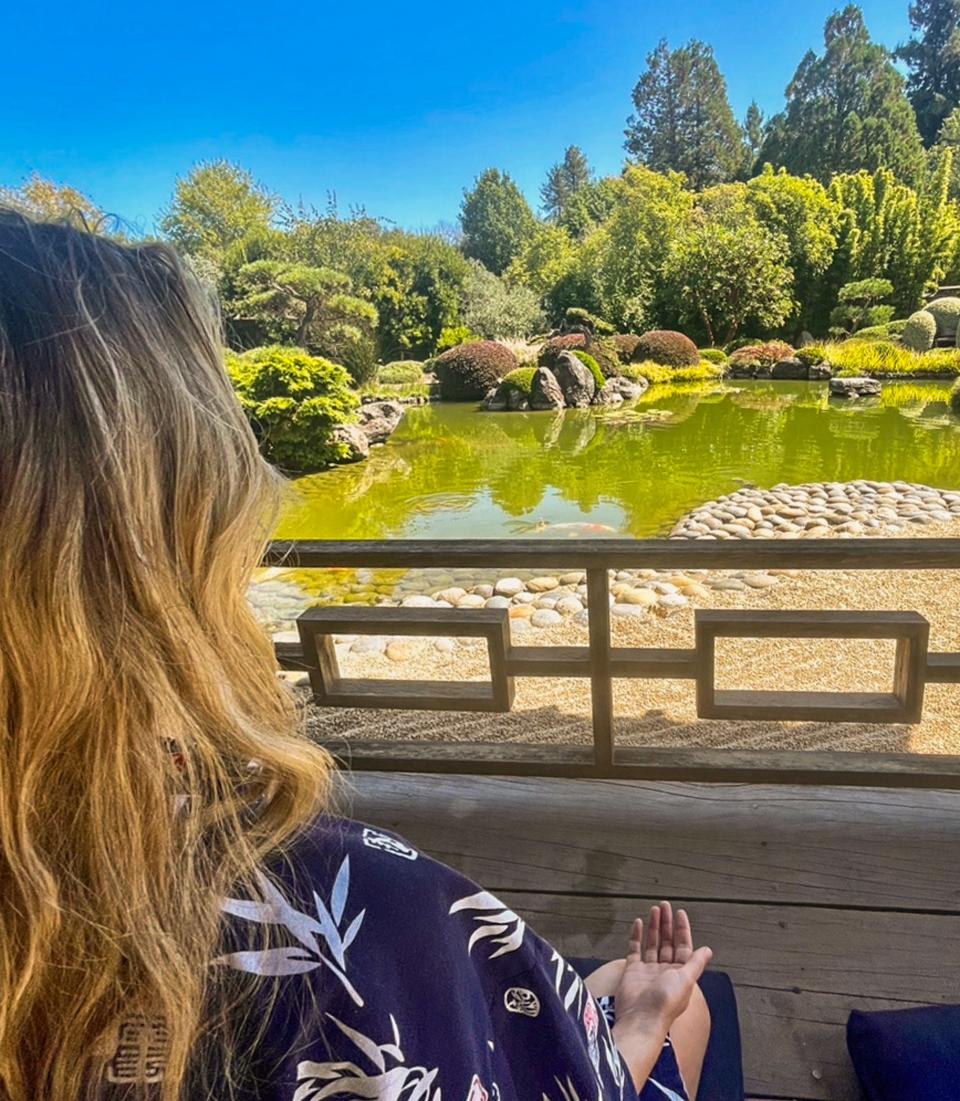 On the left side of the image, a person with long, blonde hair sits facing away from the camera with her right palm on her knee facing up. In front of her is a pond with lush bushes, hedges, and trees behind it. The sky is clear and blue.