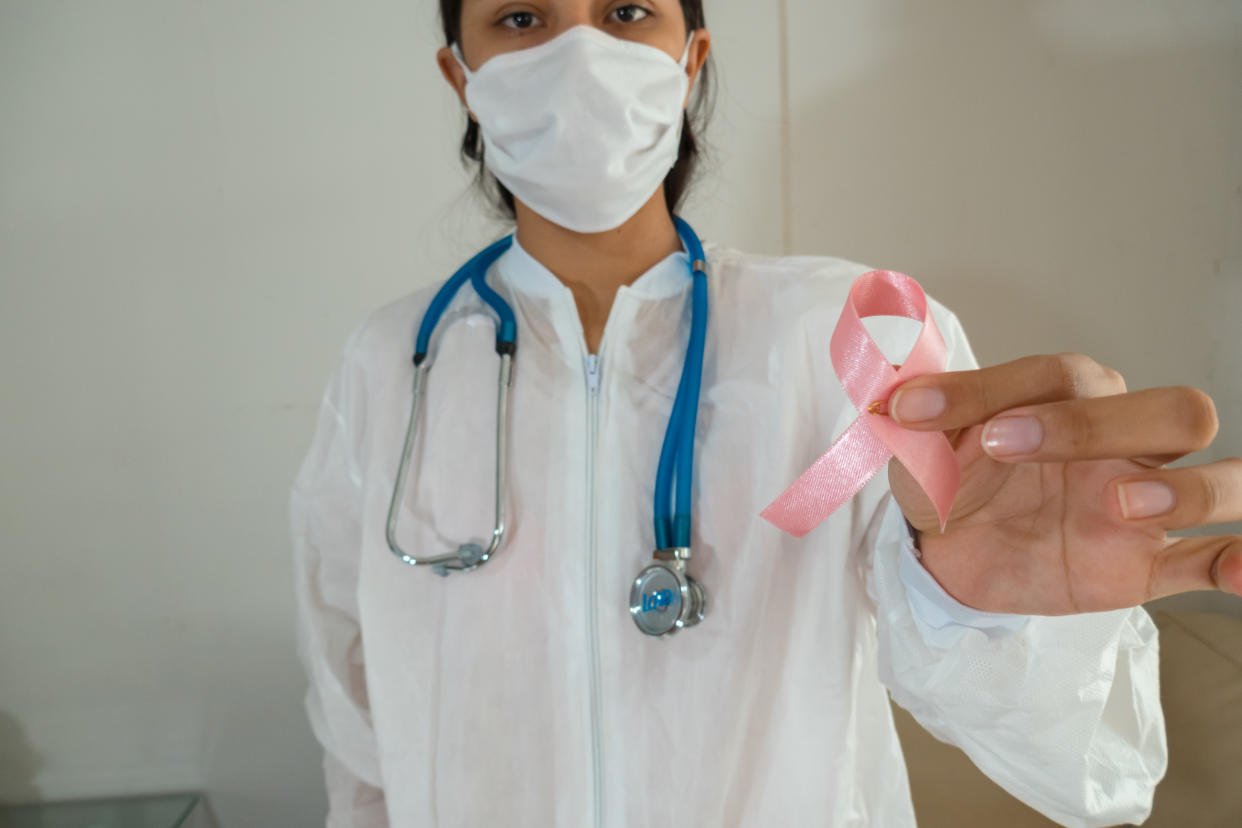 Medical worker in white scrubs and face mask with blue stethoscope around neck holds out pink ribbon