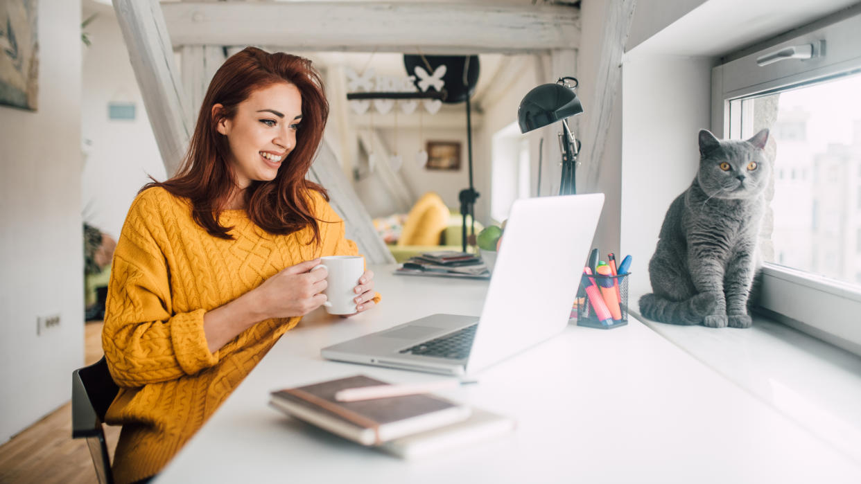 Smiling young redhead holding a cup of coffee while working on her laptop in her home office with a cat sitting in a window.