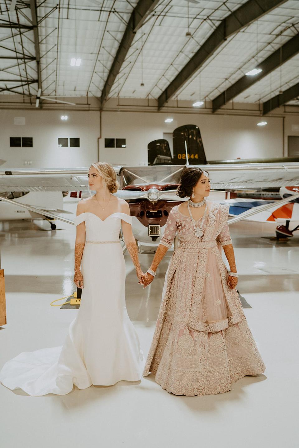 Two brides hold hands in their wedding attire and look around a museum full of airplanes.