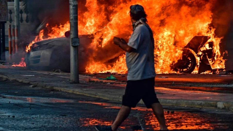 Carro en fuego y manifestante en primer plano.