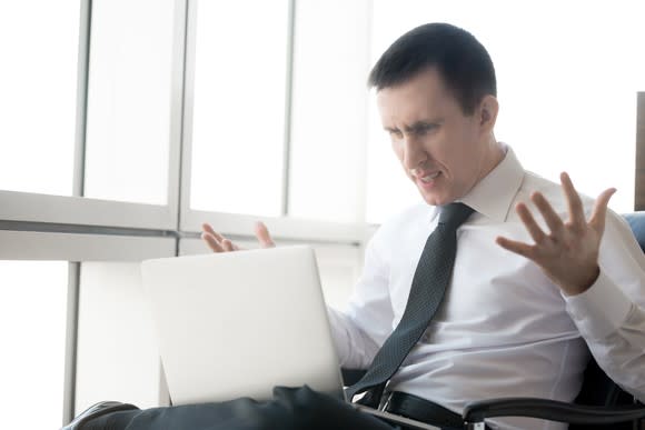 A frustrated investor throwing his hands up in the air while looking at material on his laptop.