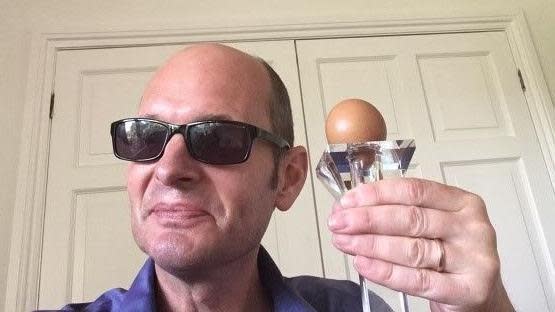 Ed has a bald head and is wearing sunglasses with black frames and holds round-shaped egg on a transparent stand in his left hand up close to his face as he smiles towards the camera.