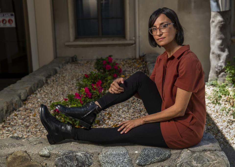 Carribean Fragoza, wearing a rust-colored shirt and black pants, sits on a stone ledge in a garden.