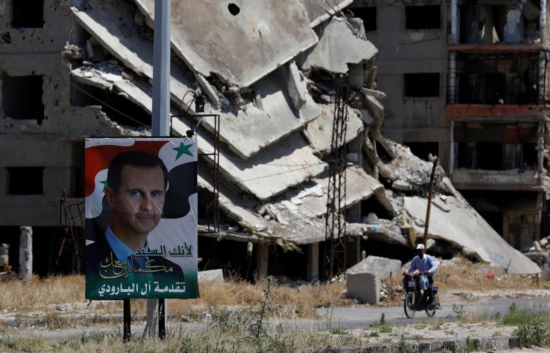 FILE PHOTO: A poster depciting Syria's President Bashar al-Assad is pictured in front of damaged buildings in Homs