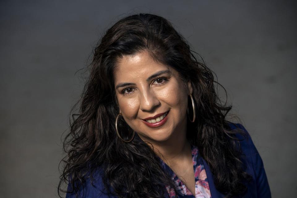 Ana-Christina Ramón, a UCLA scholar, is seen in a blue jacket with a patterned shirt, against an obscured background.