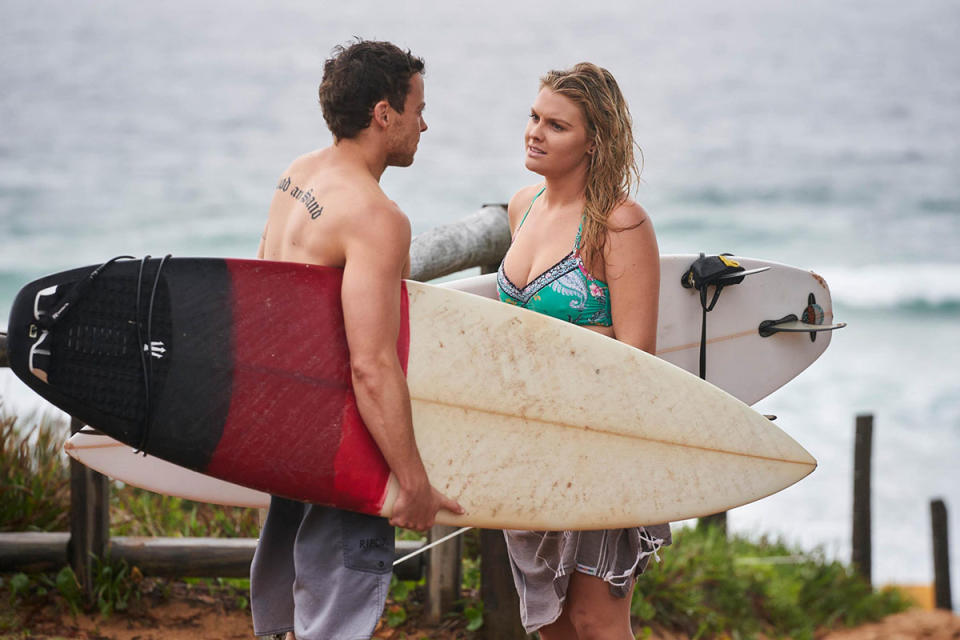 Patrick O'Connor as Dean Thompson and Sophie Dillman as Ziggy Astoni look at each other while holding surf boards