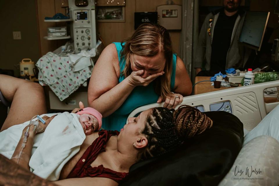 A woman on a hospital bed holds a newborn, another woman stands by emotionally, and a person observes from behind