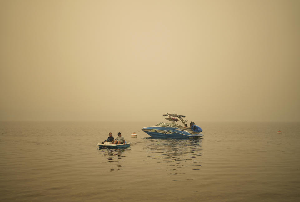 Smoke fills the air as Pat Manzuik and her husband Trevor use a paddleboat to get to shore after being given a boat ride by good samaritan Christy Dewalt, back right, back to their home they were evacuated from due to the Lower East Adams Lake wildfire, in Scotch Creek, Canada, Sunday, Aug. 20, 2023. (Darryl Dyck/The Canadian Press via AP)