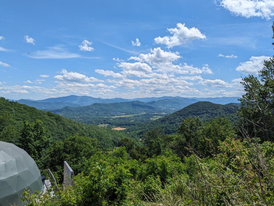 a large black piece of fiberglass covered with bolts and metal plates is on the ground next to a path leading into a forest.  rolling mountains can be seen in the distance