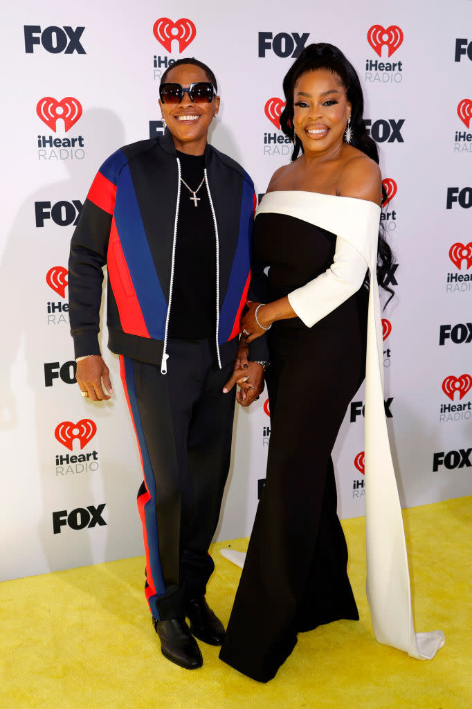 Two celebrities posing together at an event, one in a stylish track suit and the other in an elegant off-the-shoulder gown