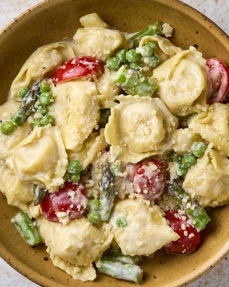 overhead shot of creamy tortellini primavera in a small yellow bowl, topped with cheese