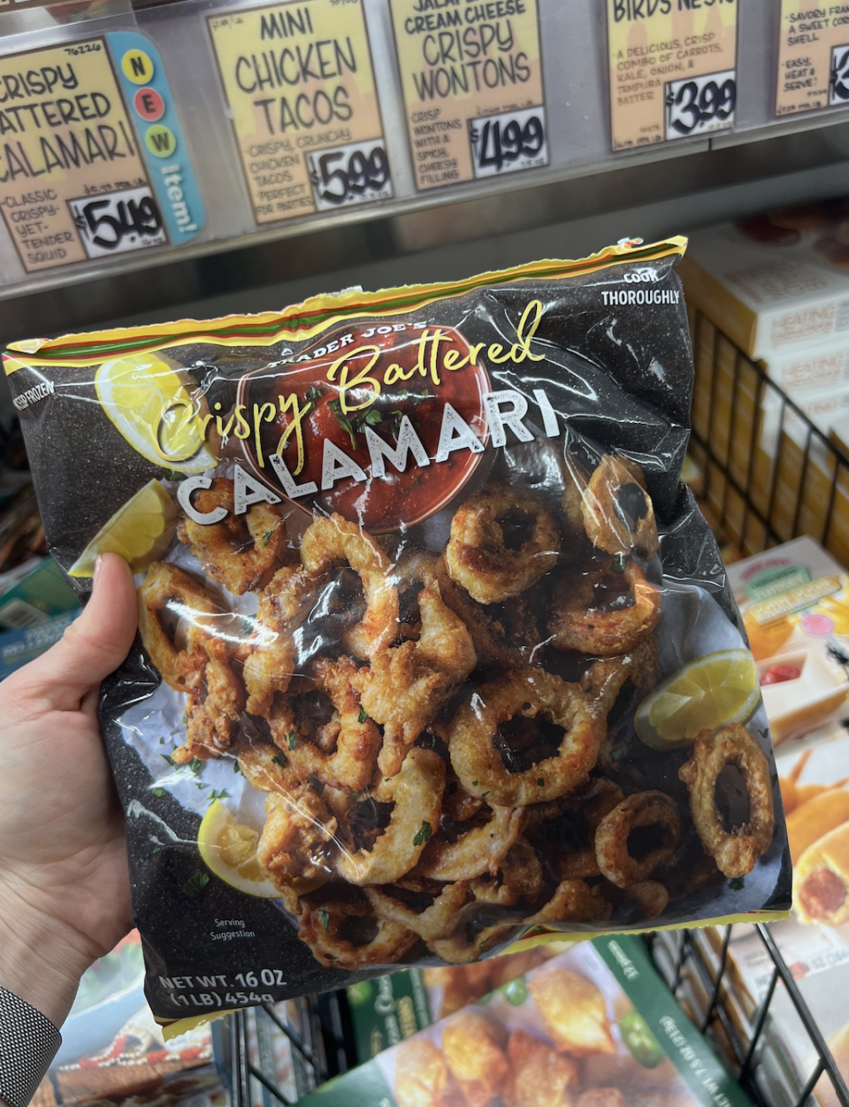 Hand holding Trader Joe's Crispy Battered Calamari package in a grocery store frozen food section with other food items visible in the background