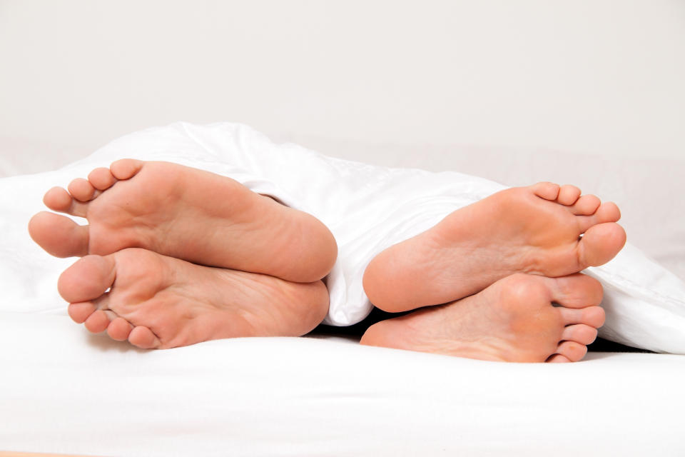 Two pairs of feet stick out from under a blanket on a bed, facing away from each other