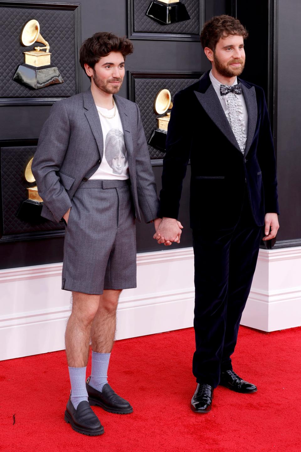 Noah in a dark grey blazer with matching high-waisted Bermuda shorts. He's wearing a white T-shirt with a screenprint of Barbra Streisand's face. He's wearing light blue dress socks and loafers. Benn is in a dark navy velvet suit.