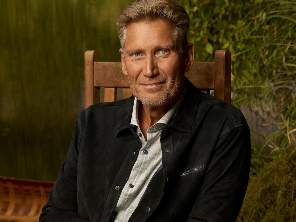 Gerry Turner poses for photos in a wooden chair wearing a black jacket and jeans.