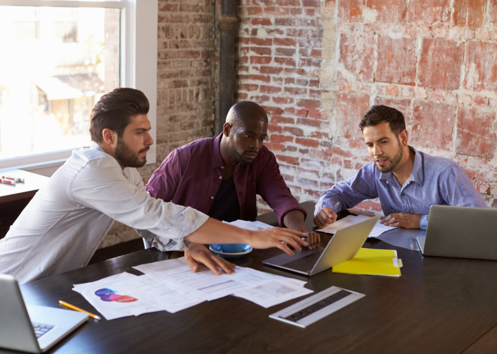 Three people collaborating sit at a desk with three laptops and papers spread out in front of them. 