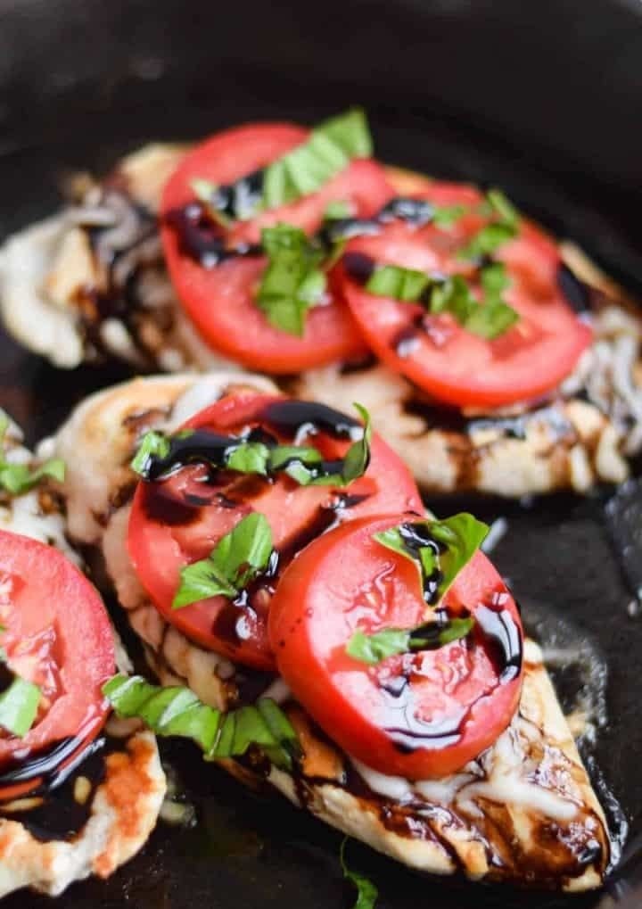 Three chicken breasts topped with tomato, cheese, basil, and balsamic drizzle in a skillet