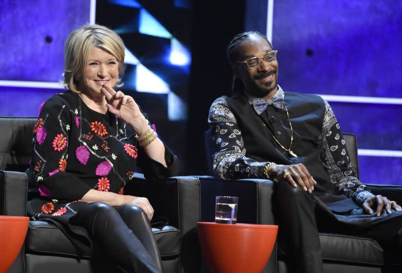 Martha Stewart in floral blouse and Snoop Dogg in vest and paisley bandana shirt smile while sitting on leather chairs.