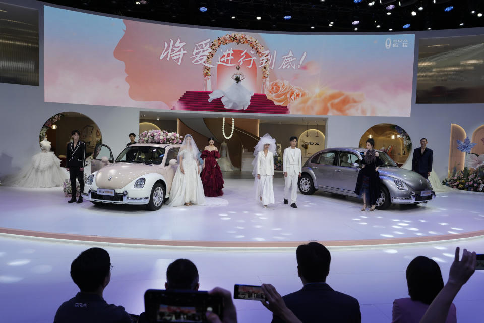 Visitors film models in wedding gowns showcasing the Ora, a Chinese-made electric car brand during the Auto Shanghai 2023 at the National Exhibition and Convention Center in Shanghai, China, Tuesday, April 18, 2023. Global and Chinese automakers plan to unveil more than a dozen new electric SUVs, sedans and muscle cars this week at the Shanghai auto show, their first full-scale sales event in four years in a market that has become a workshop for developing electrics, self-driving cars and other technology. (AP Photo/Ng Han Guan)