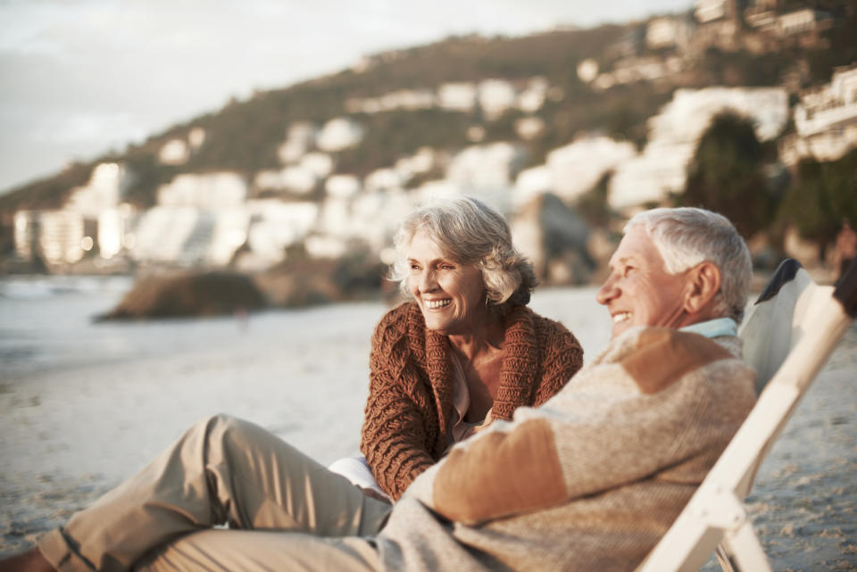 An elderly couple, both in cozy sweaters, enjoy a relaxing time on the beach, sharing a moment of laughter with blurry distant buildings in the background