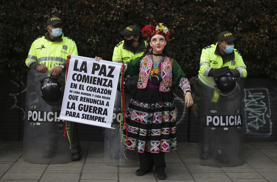 A woman dressed in a traditional Mexican dress and mask holds a sign that reads in Spanish "Peace begins in the heart of every man and woman to renounce war forever." as police stand guard on the sidelines of an anti-government march in Bogota, Colombia, Wednesday, May 19, 2021. Colombians have taken to the streets for weeks across the country after the government proposed tax increases on public services, fuel, wages and pensions. (AP Photo/Ivan Valencia)