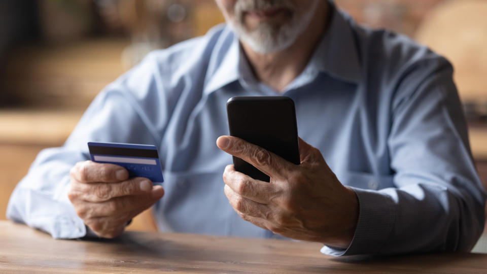 Close up mature man using phone, holding plastic credit or debit card, senior grey haired customer making secure internet payment, shopping or browsing online banking service, entering information