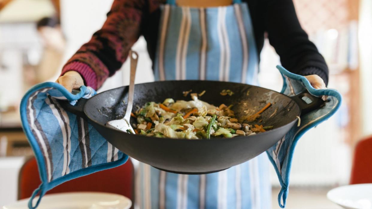  A person holding a wok full of healthy stir fry vegetables with oven gloves. 