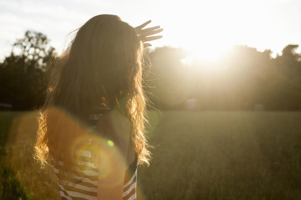 A person with long hair wearing a striped top stands with their back to the camera, shielding their eyes from the sun, while gazing into a vast field