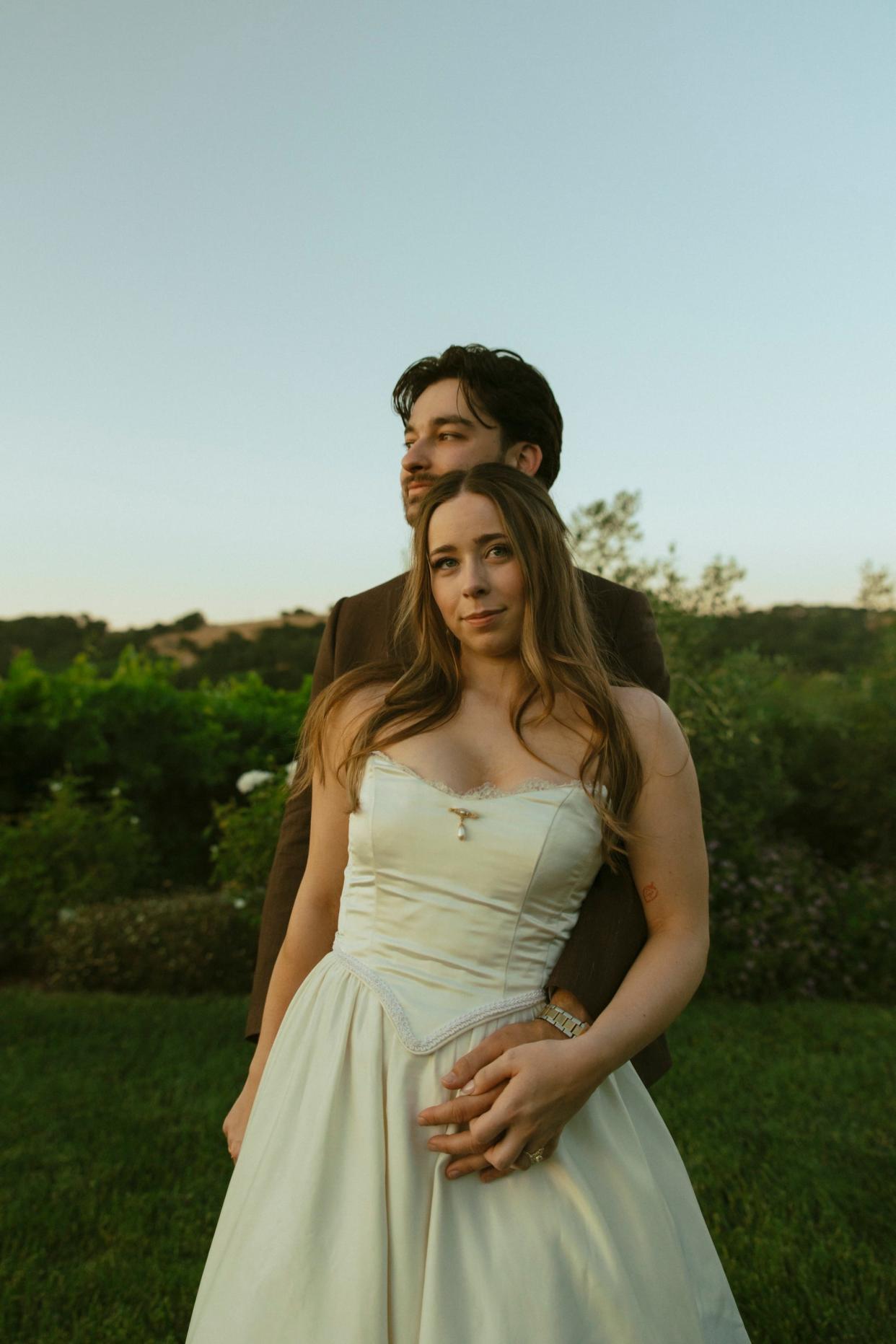 A groom embraces his bride from behind in a field.