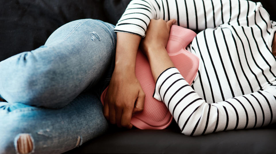 Person lying down, clutching a hot water bottle to their abdomen, wearing a striped top and jeans