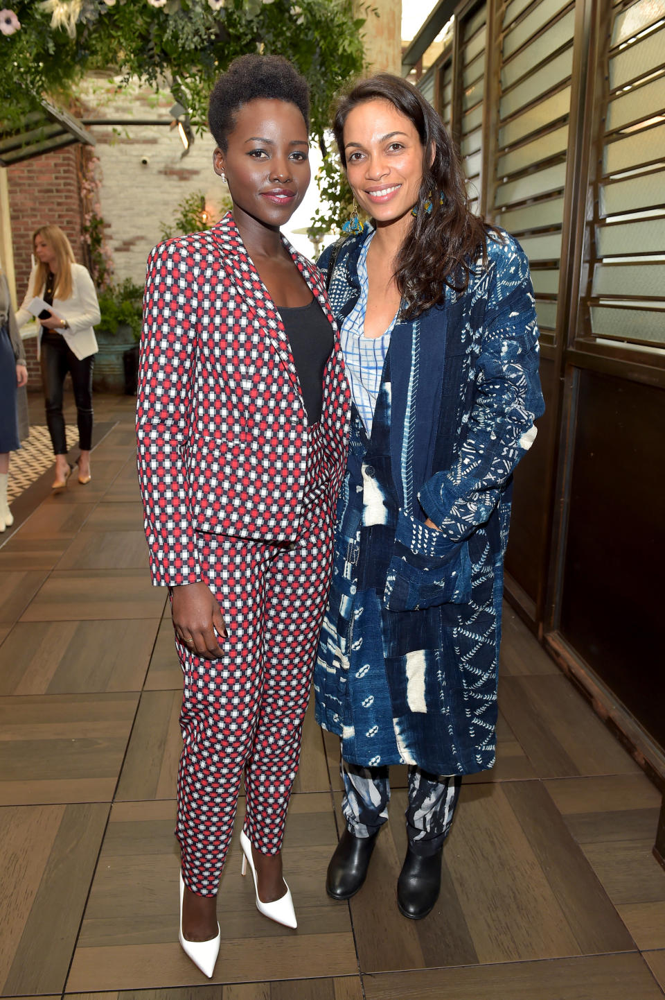 Actors Lupita Nyong'o and Rosario Dawson wearing suits - Lupita in a white suit with bold red and black geometric patterns, and Rosario in a denim blue pattern-clash combo.