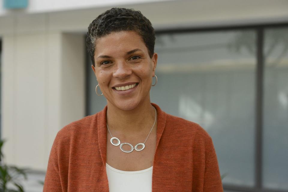 michelle alexander smiles at the camera while standing outside, she wears an orange sweater over a white top, a silver necklace and hoop earrings