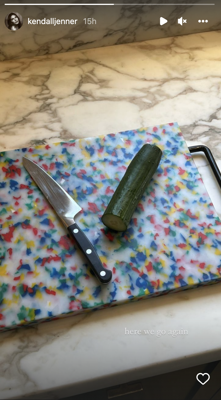 An oversized cutting board holds a large knife on the left with a partially chopped cucumber next to it