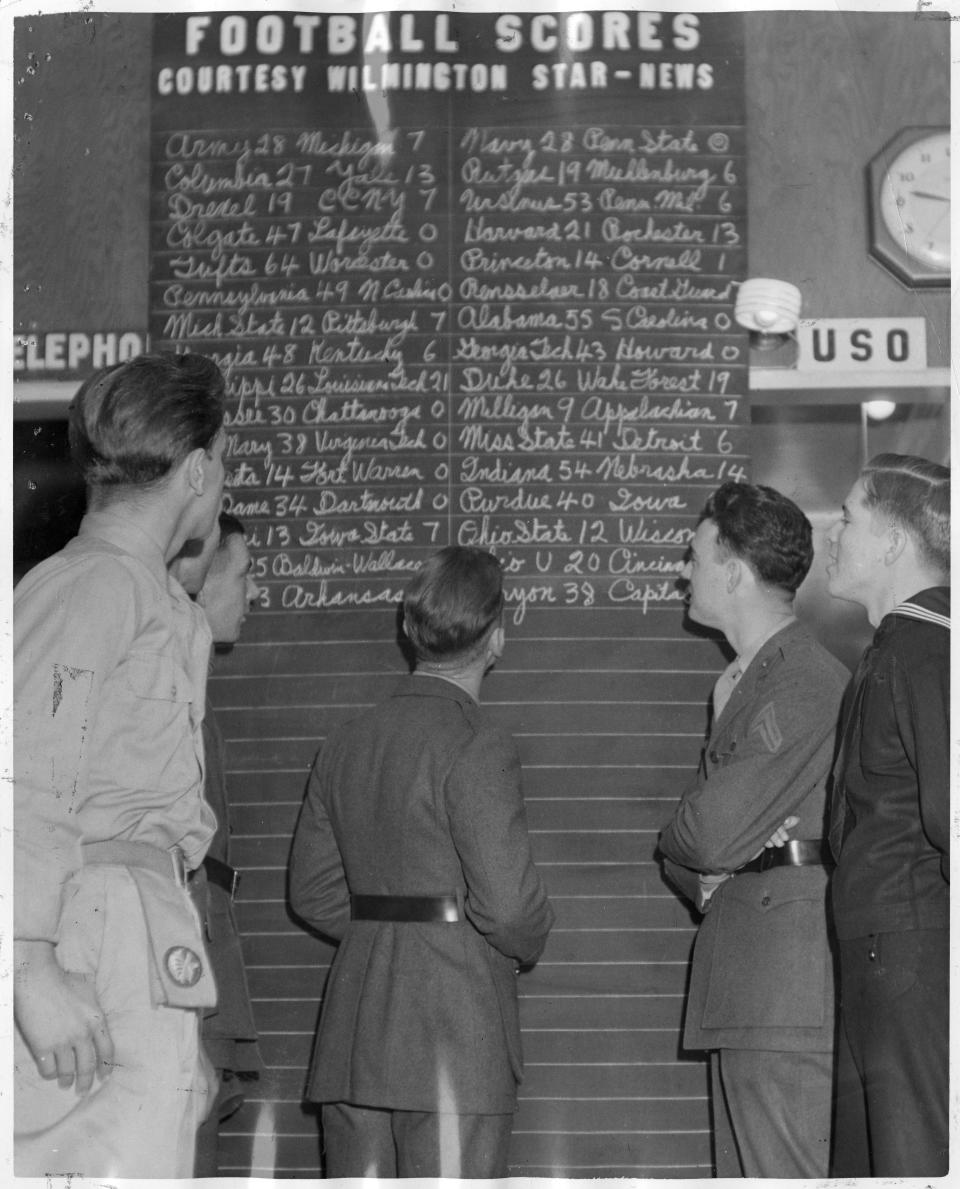 Servicemen look for home town football scores as they are added as received every Saturday night.  The large ruled chalkboard reads “FOOTBALL SCORES/COURTESY WILMINGTON STAR-NEWS,” and was propped against the lobby wall by the door leading to the switchboard and telephone booths. Two Marines, a sailor and an Army paratrooper are reading the scores.  The picture is captioned “October 1945.”  The exact date is not specified.