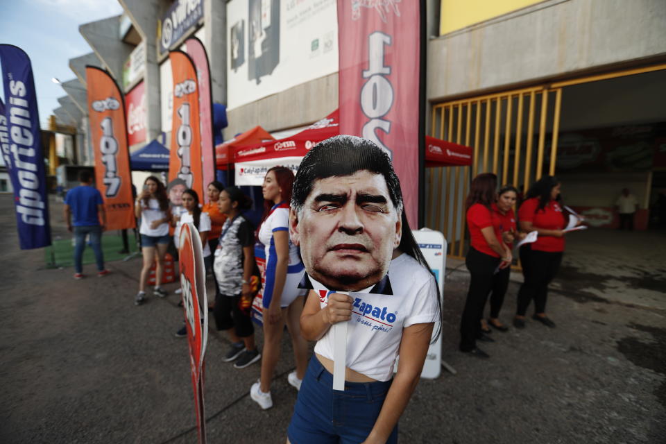 An event promoter holds a mask of Diego Maradona, head coach of Dorados de Sinaloa soccer team, at the venue of a second-tier national league soccer match against Cafetaleros, his first game as coach for Dorados in Culiacan, Mexico, Monday, Sept. 17, 2018. (AP Photo/Eduardo Verdugo)