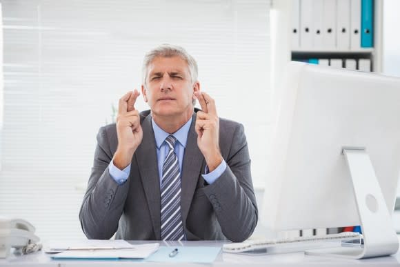 Businessman sitting at desk with computer, crossing his fingers hard with his eyes closed.
