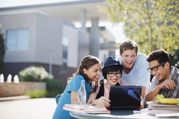 College-age people gathered around a laptop computer on a table outdoors.