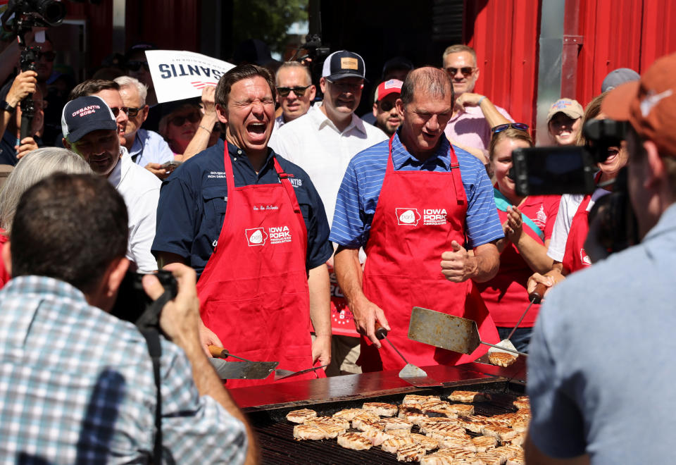 Ron DeSantis, wearing a red apron and appearing to wince with his eyes squeezed closed, holds a spatula in front of an outdoor grill among other people, many of whom are also wearing red aprons.